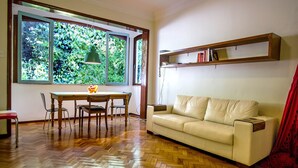 A room with a wooden herringbone floor, a beige sofa, a dining table with chairs, and a window with a view of greenery.