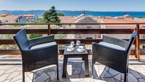 A balcony with wicker chairs and a table, overlooking a coastal town with red-roofed buildings and the sea.