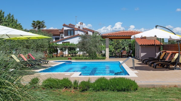 A swimming pool with lounge chairs and umbrellas, surrounded by greenery and a building with a tiled roof.