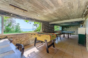 A foosball table in a room with a view of the sea, a stone wall, and a green table.