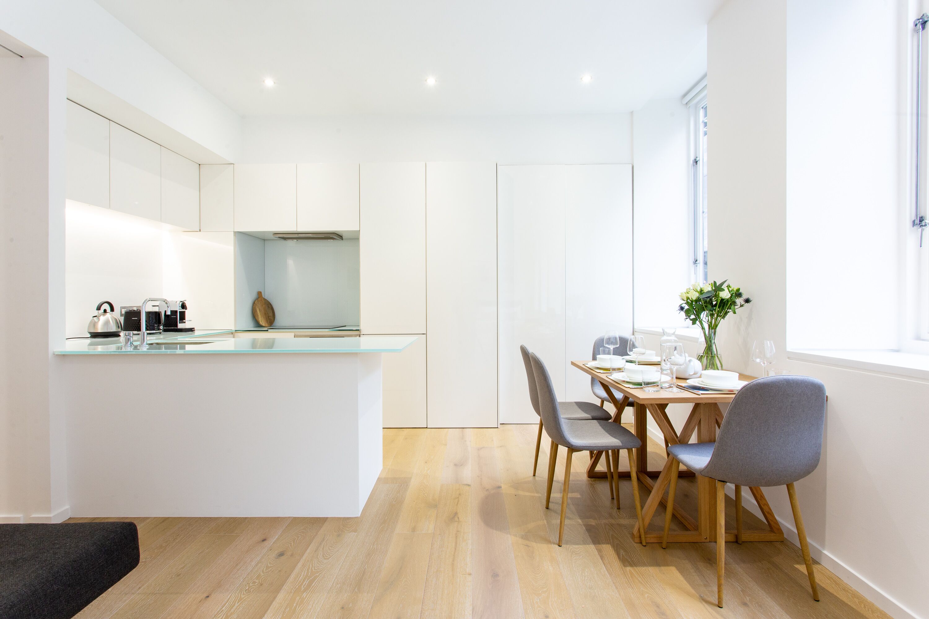 Une cuisine moderne avec des meubles blancs, un îlot central en verre et un coin repas comprenant des chaises et une table en bois.