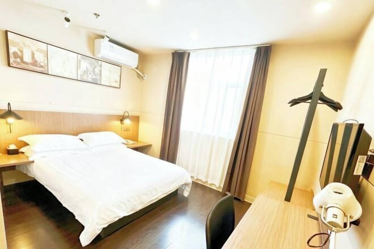 A hotel room with a bed, bedside tables, a desk, a chair, a hanging coat rack, and a window with curtains.