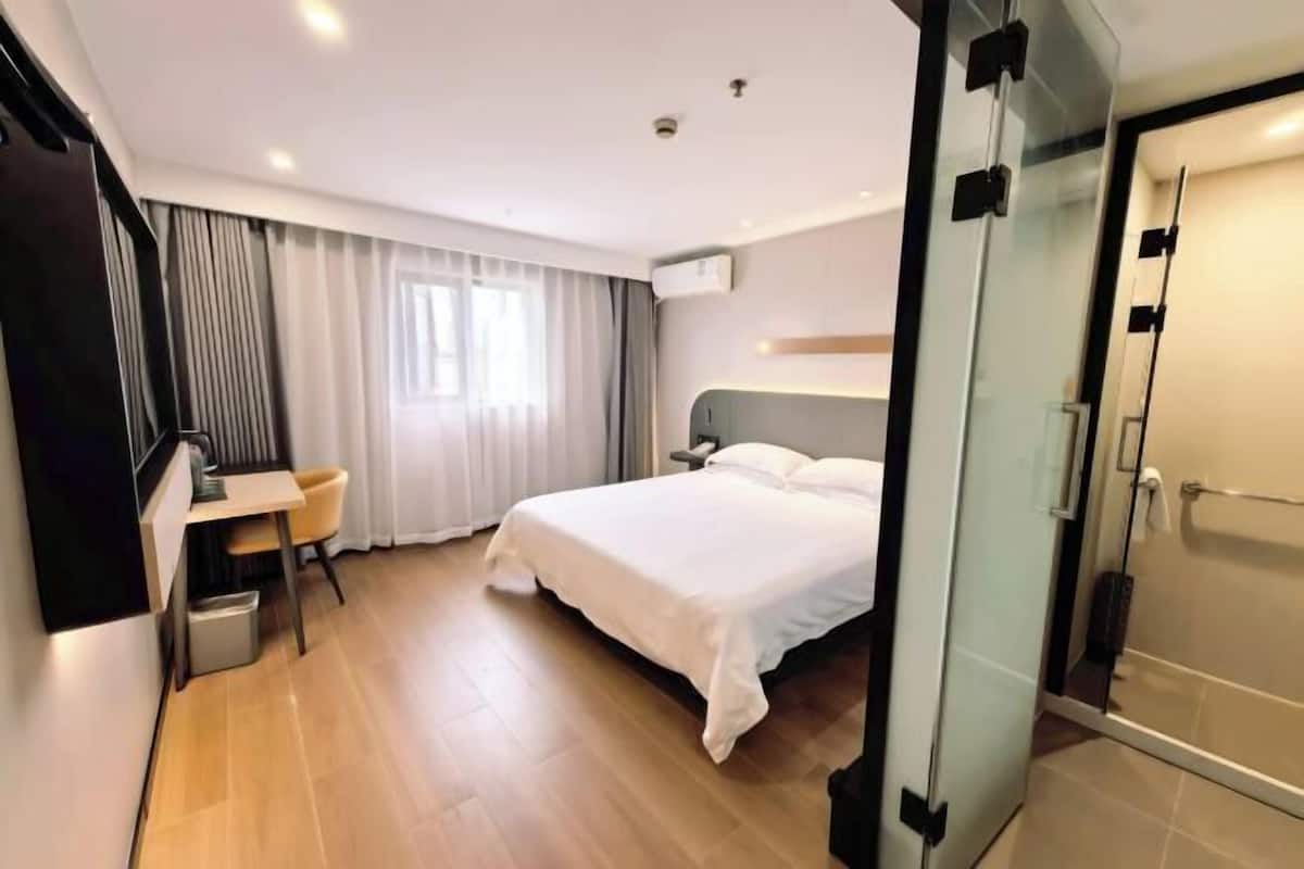 A hotel room with a bed, a desk, a chair, a shower area, and a window with curtains.