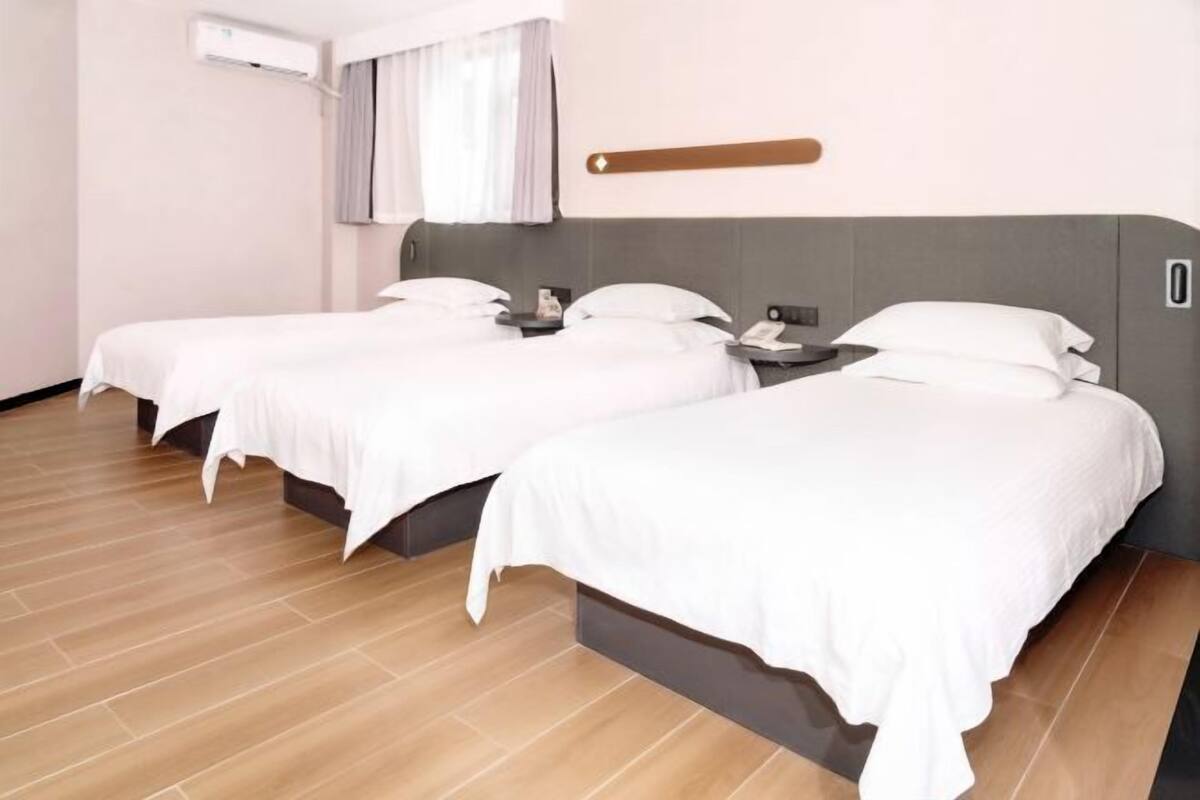 A hotel room with two single beds, a bedside table with a lamp, an air conditioning unit, and a window with curtains.