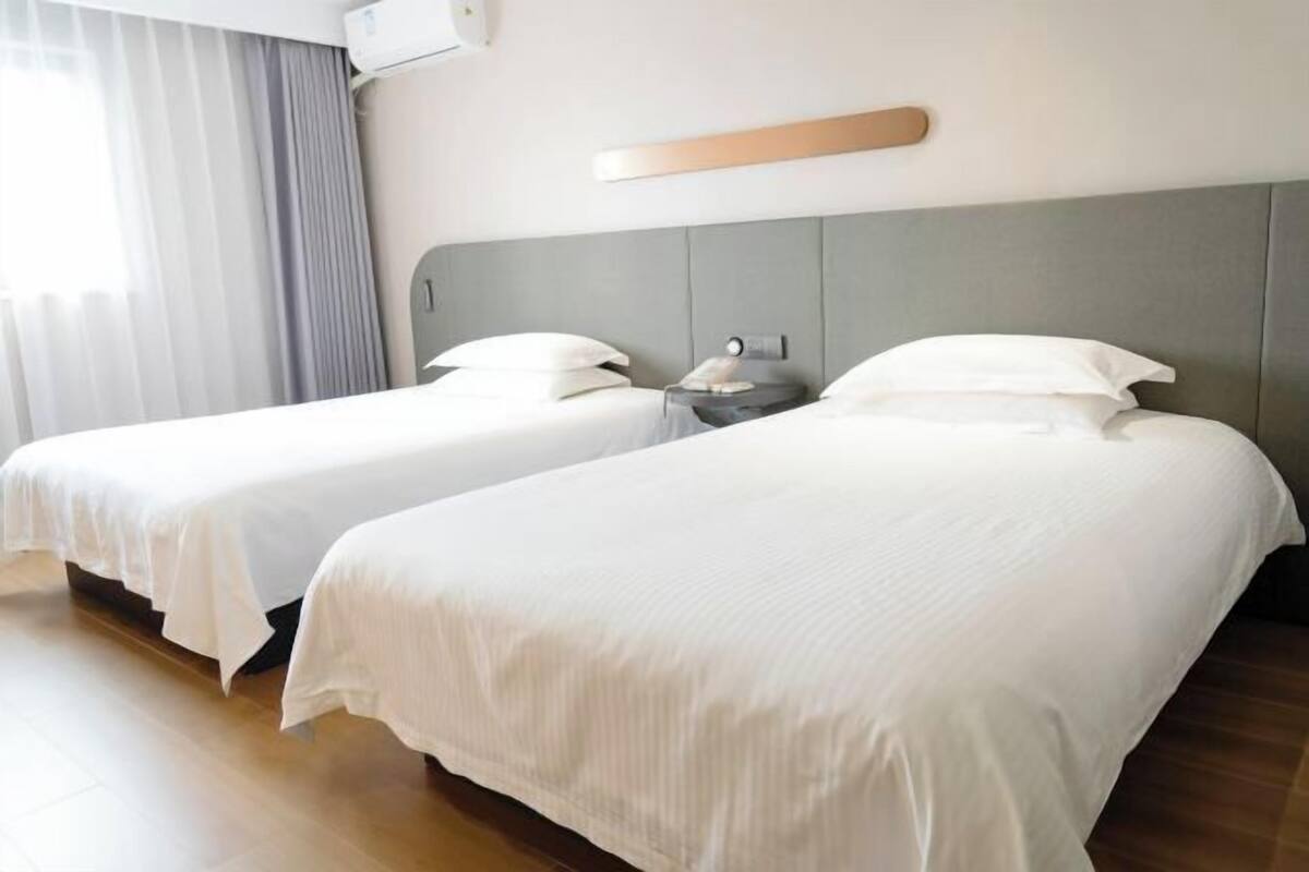 A hotel room with two beds, a bedside table with a lamp, a wall-mounted light, and a window with curtains.
