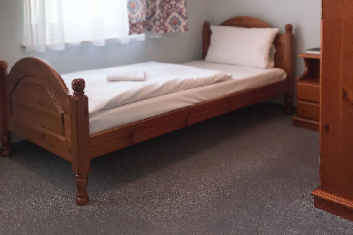 A single bed with a wooden headboard in a room with a carpeted floor, a window with patterned curtains, and a framed picture on the wall.