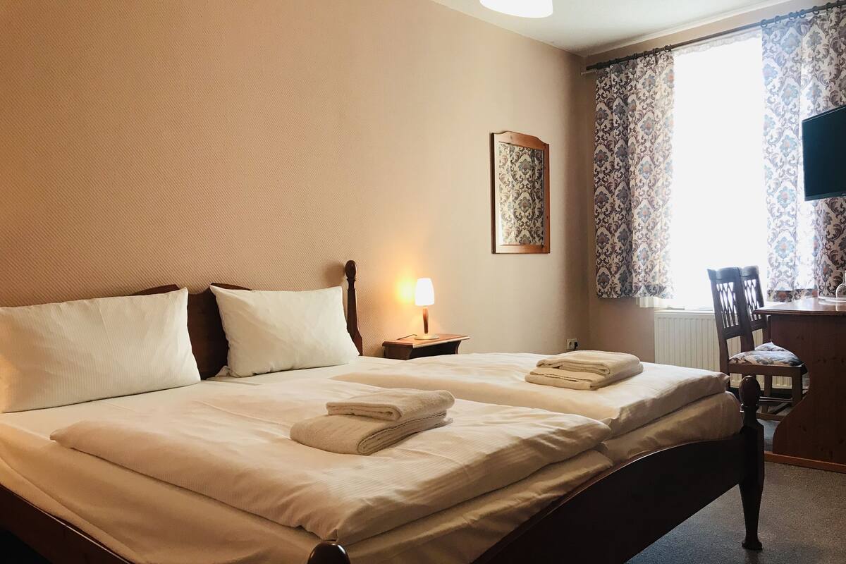 A neatly made bed with white linens and two folded towels, a bedside lamp, a framed artwork on the wall, and a window with patterned curtains.