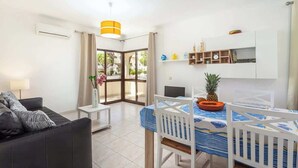 Un saló modern amb un sofà, una taula de dinar amb cadires, una taula blanca petita i una zona de cuina amb nevera, prestatges i una làmpada penjant.