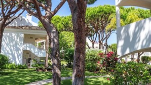 Un camí pavimentat que condueix a un edifici amb façana blanca i un jardí ple d'arbres i arbustos.
