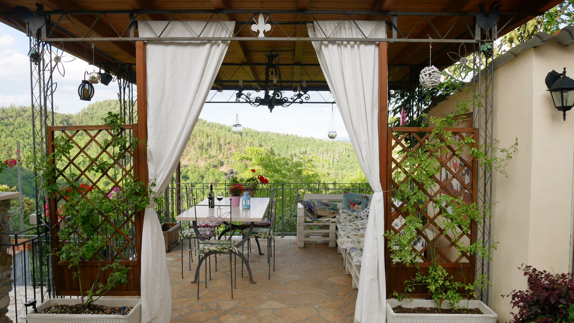 Een overdekte terras met een tafel en stoelen, omringd door potplanten en een prachtig uitzicht op heuvels.