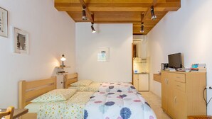 Ein Schlafzimmer mit einem hölzernen Bett, einem Nachttisch mit Lampe, einer Holzbank, einem Fernseher auf einem Schrank und einer Küchenzeile im Hintergrund.