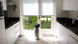 Une cuisine avec des meubles blancs, une poubelle en acier inoxydable et une fenêtre avec des stores.