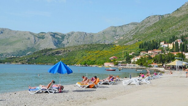 Una playa con gente tomando el sol, una sombrilla azul y montañas al fondo.