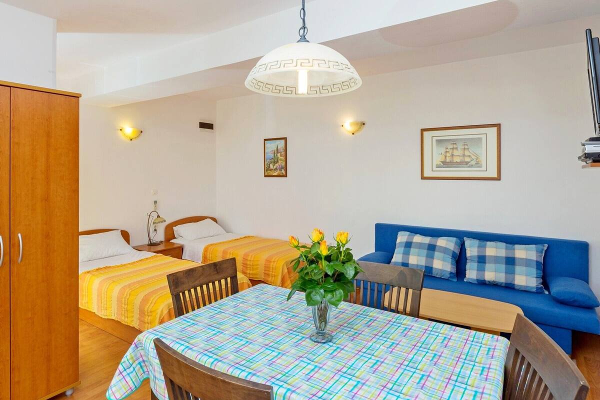 A room with a dining table, two beds, a blue sofa, and a hanging light fixture.