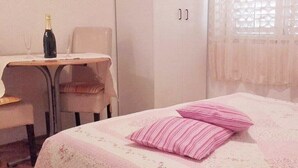 A room with a bed, a chair, a table with a bottle and glass, and a window with lace curtains. - Apartments Jela, (15070), Makarska, hr (Makarska)