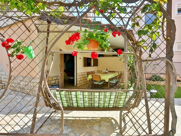 A cozy outdoor seating area with a swing chair, a table, and chairs, surrounded by a metal fence and greenery. - Apartments Ivan, (15798), Solin, hr (Solin)