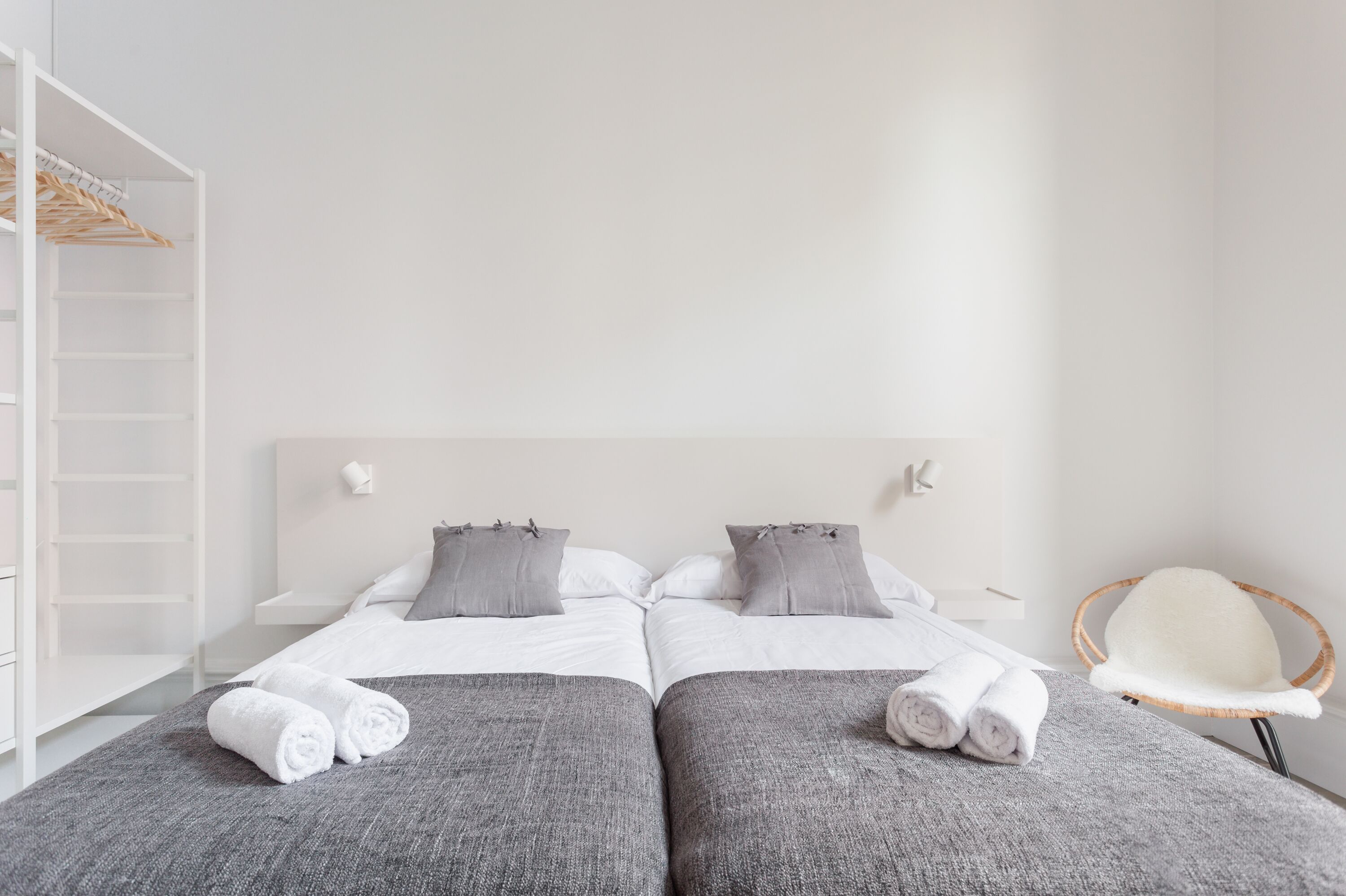 Una cama doble con ropa de cama y almohadas grises, un armario blanco y una silla de mimbre.