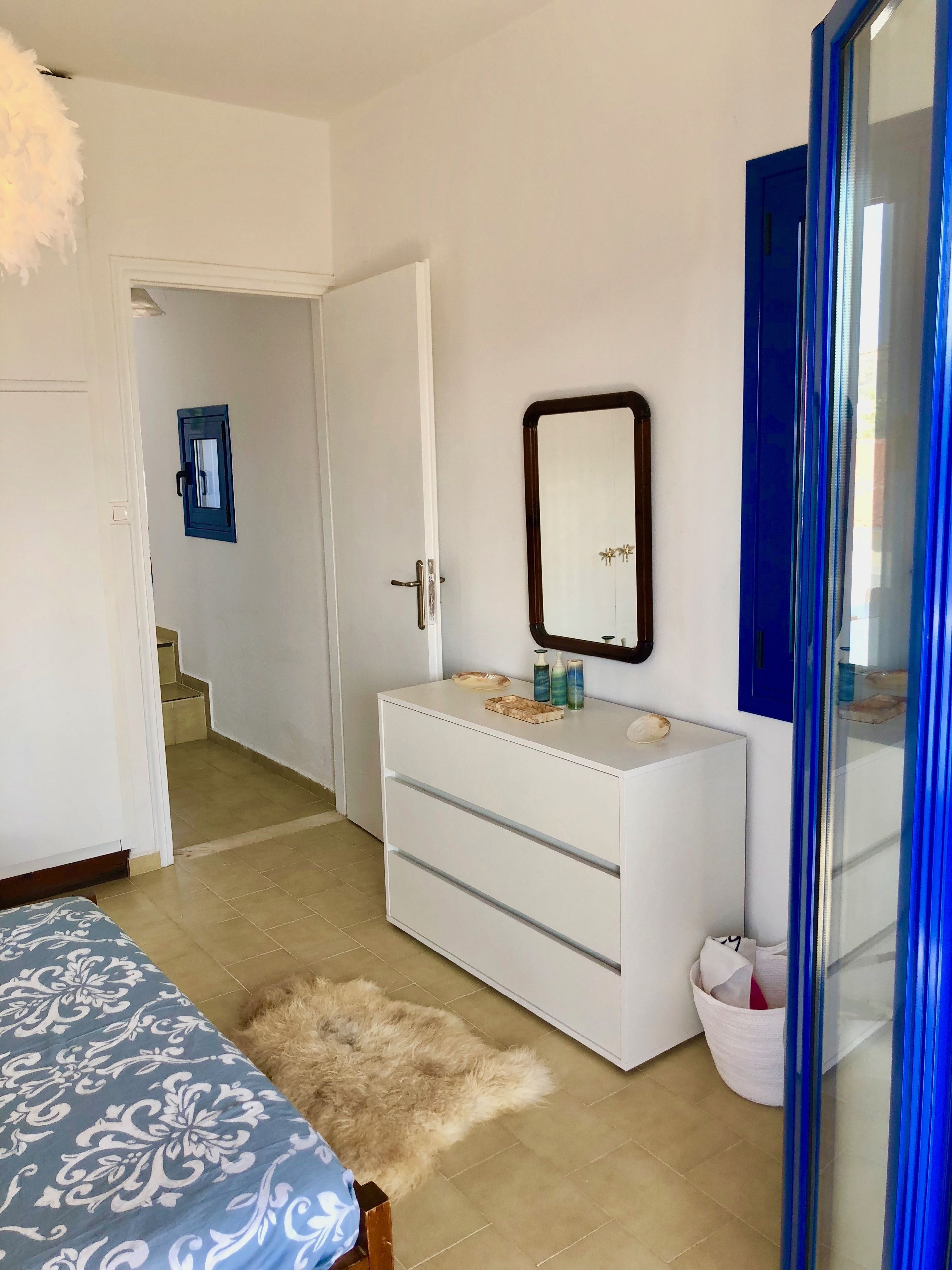 A bedroom with a white dresser, a mirror, a fluffy rug, and a bed with a patterned bedspread.