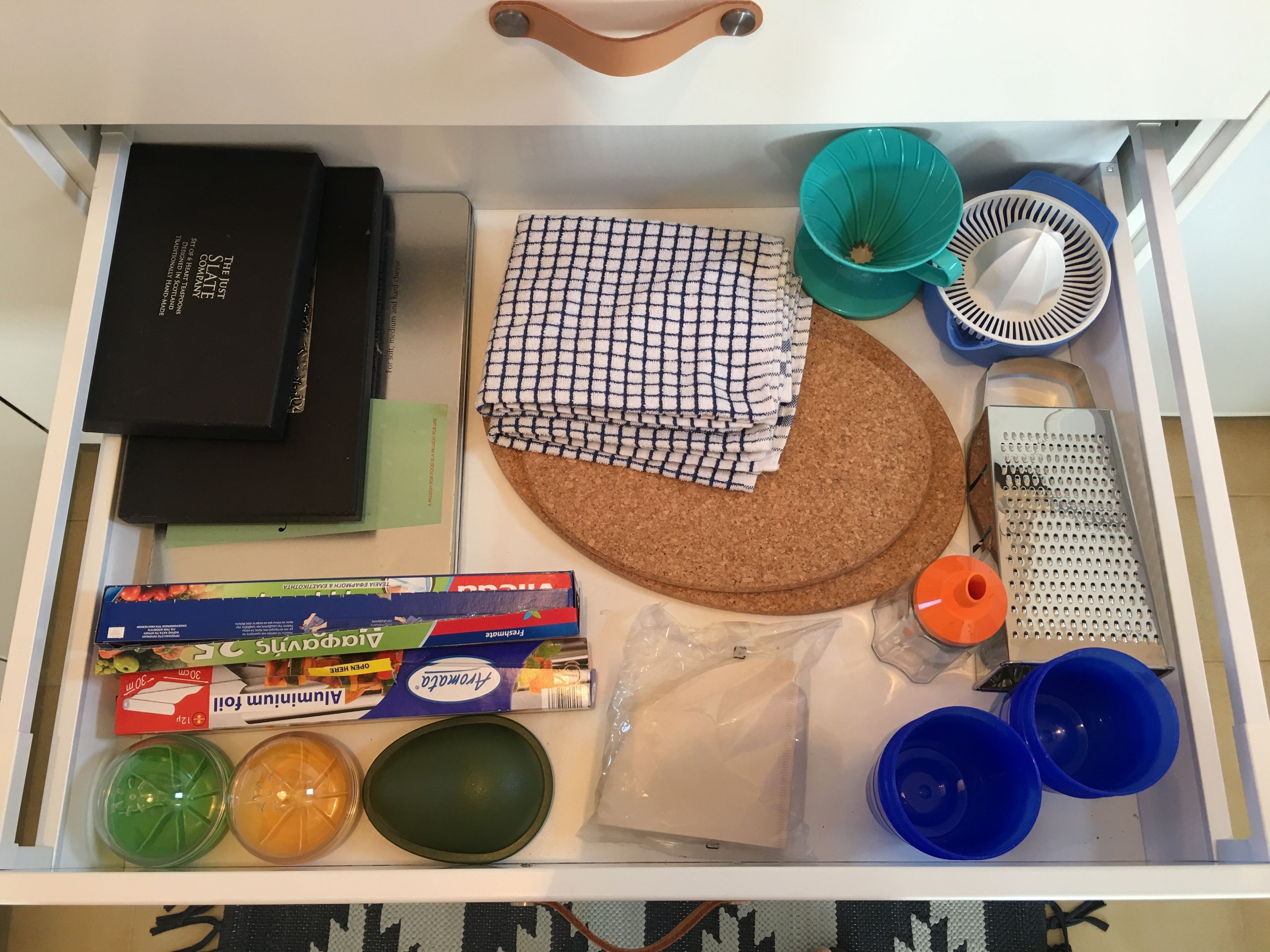 A drawer containing various items including a cork mat, a checkered cloth, a grater, plastic containers, and a box of Airwaves.