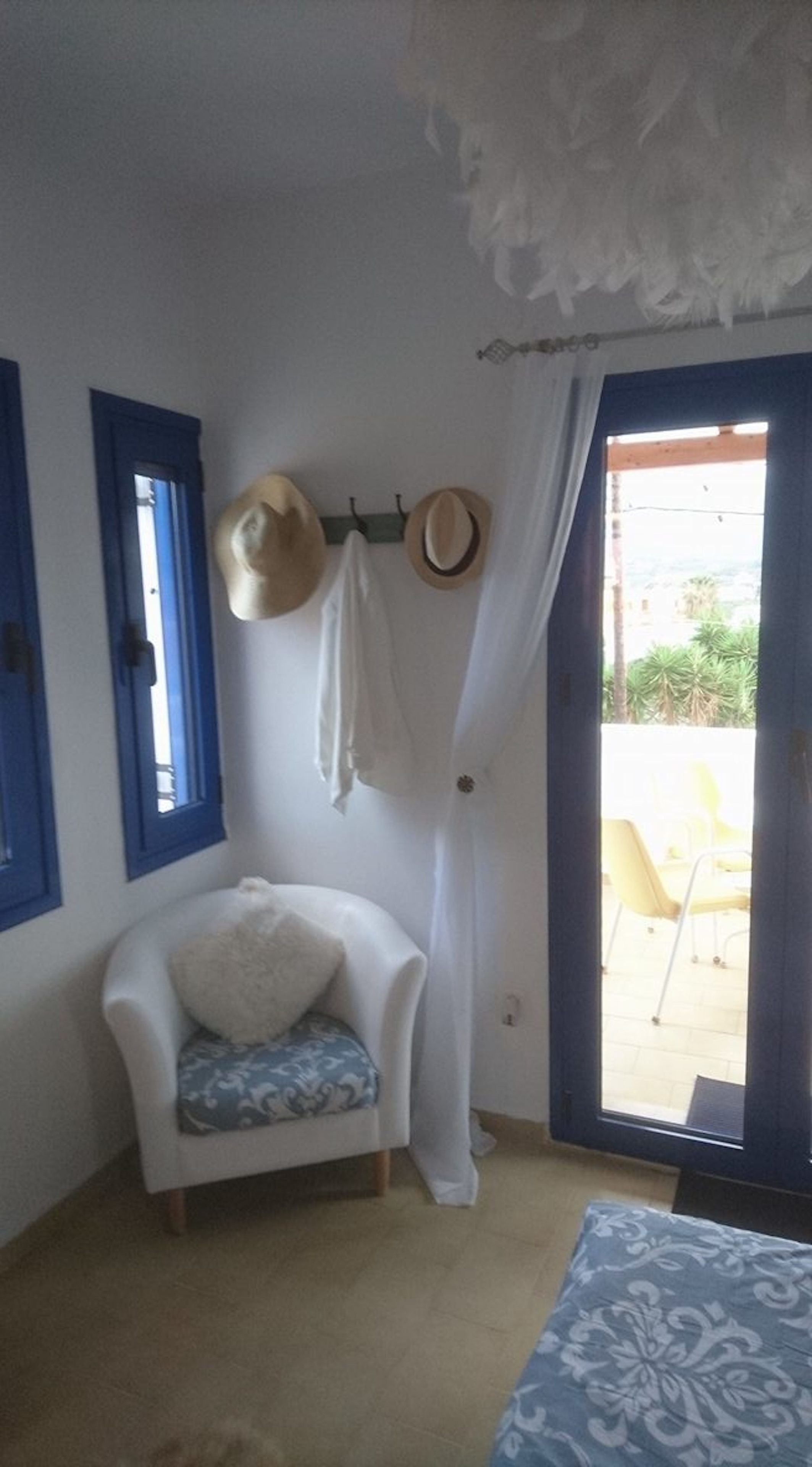 A room with a white armchair, a small table, a hat rack with hats, and a view of an outdoor area with a yellow chair.