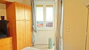 Een slaapkamer met een bed, een houten kledingkast, een televisie, een raam met uitzicht op gebouwen en een vaas met takken.