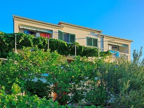 Stands amidst lush greenery, featuring a simple two-story design with a flat roof and upper-floor balcony.