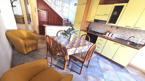 Een compacte keuken met gele kastjes, een eettafel met een tafelkleed met dessin, en een gele fauteuil.