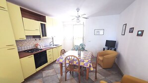 Een compacte keuken met gele kastjes, een eettafel met stoelen, een televisie en een plafondventilator.