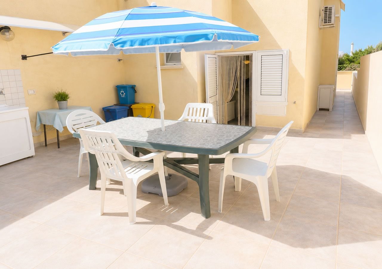 An outdoor patio with a table and chairs, a striped umbrella, and a small table with a plant.
