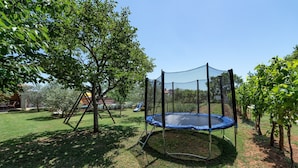 A trampoline in a backyard with a tree and a house in the background. - Apartments Neva, (15356), Štinjan, hr (Štinjan)