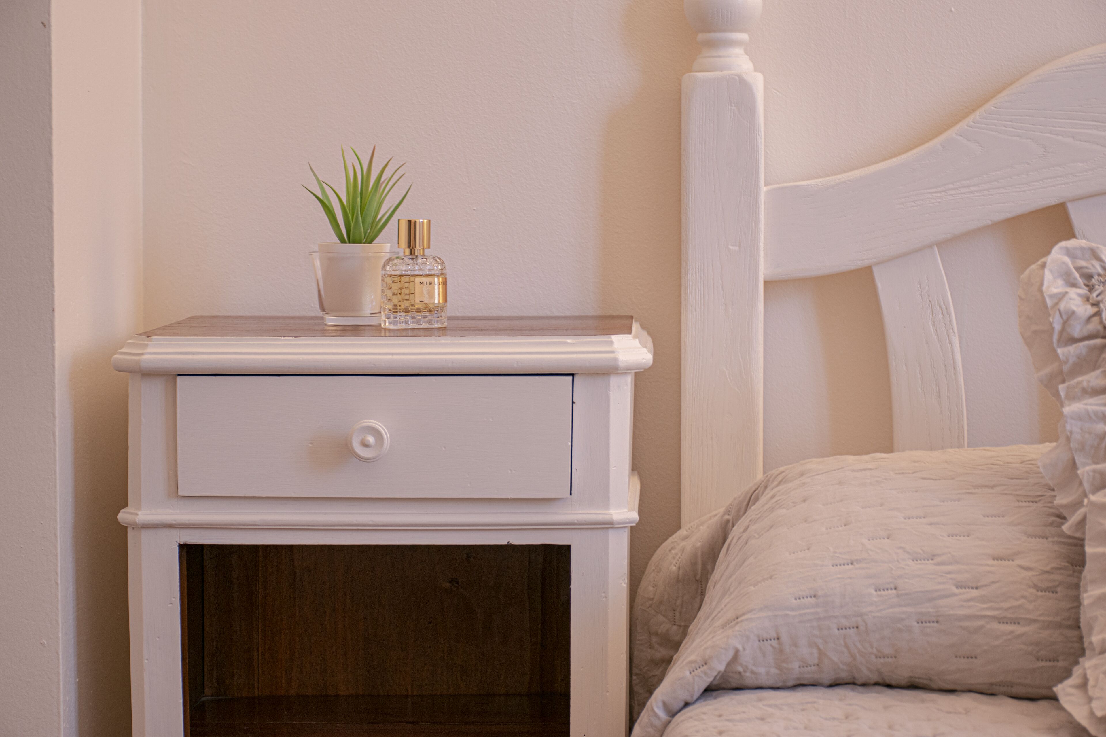 A white bedside table with a drawer, a small potted plant, and a bottle of perfume.