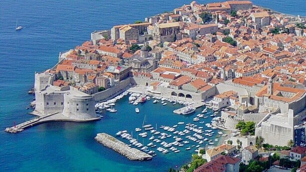 Vue aérienne d’une ville côtière dotée d’une fortification historique, d’un port où se trouvent de nombreux bateaux et d’un centre urbain dense avec des bâtiments aux toits rouges.