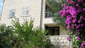 Un bâtiment à deux étages doté d’un balcon, entouré d’une végétation luxuriante et de fleurs violettes.