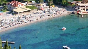Vue aérienne d’un complexe hôtelier en bord de mer, avec de nombreuses chaises longues et des parasols, un petit bateau et un bâtiment doté d’un toit rouge.