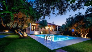 Una casa moderna con piscina, rodeada de exuberante vegetación e iluminada por luces cálidas.