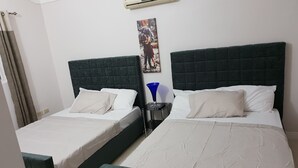 A hotel room with two beds, each with a dark headboard and white bedding. There is a wall-mounted air conditioner above the beds, a small table with a blue lamp, and a painting on the wall. - Kensington Oasis 2b/3bth- New Kingston (24 hrs security, free wifi, hot water) (New Kingston)