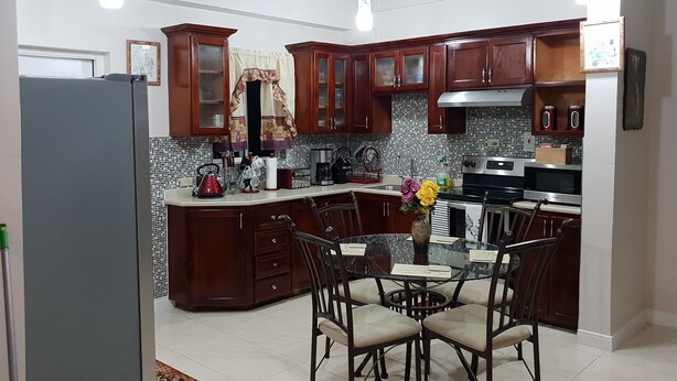 A kitchen with wooden cabinets, a tiled backsplash, a refrigerator, a stove, and a dining table with chairs. - Kensington Oasis 2b/3bth- New Kingston (24 hrs security, free wifi, hot water) (New Kingston)
