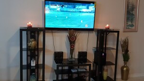 A living room with a TV mounted on a black stand, surrounded by shelves with decorative items and candles. - Kensington Oasis 2b/3bth- New Kingston (24 hrs security, free wifi, hot water) (New Kingston)