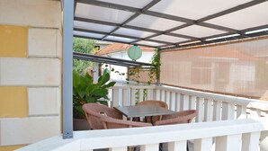 Un espace extérieur aménagé avec des meubles de jardin en osier, une table, une balustrade blanche et une vue sur une maison en arrière-plan.