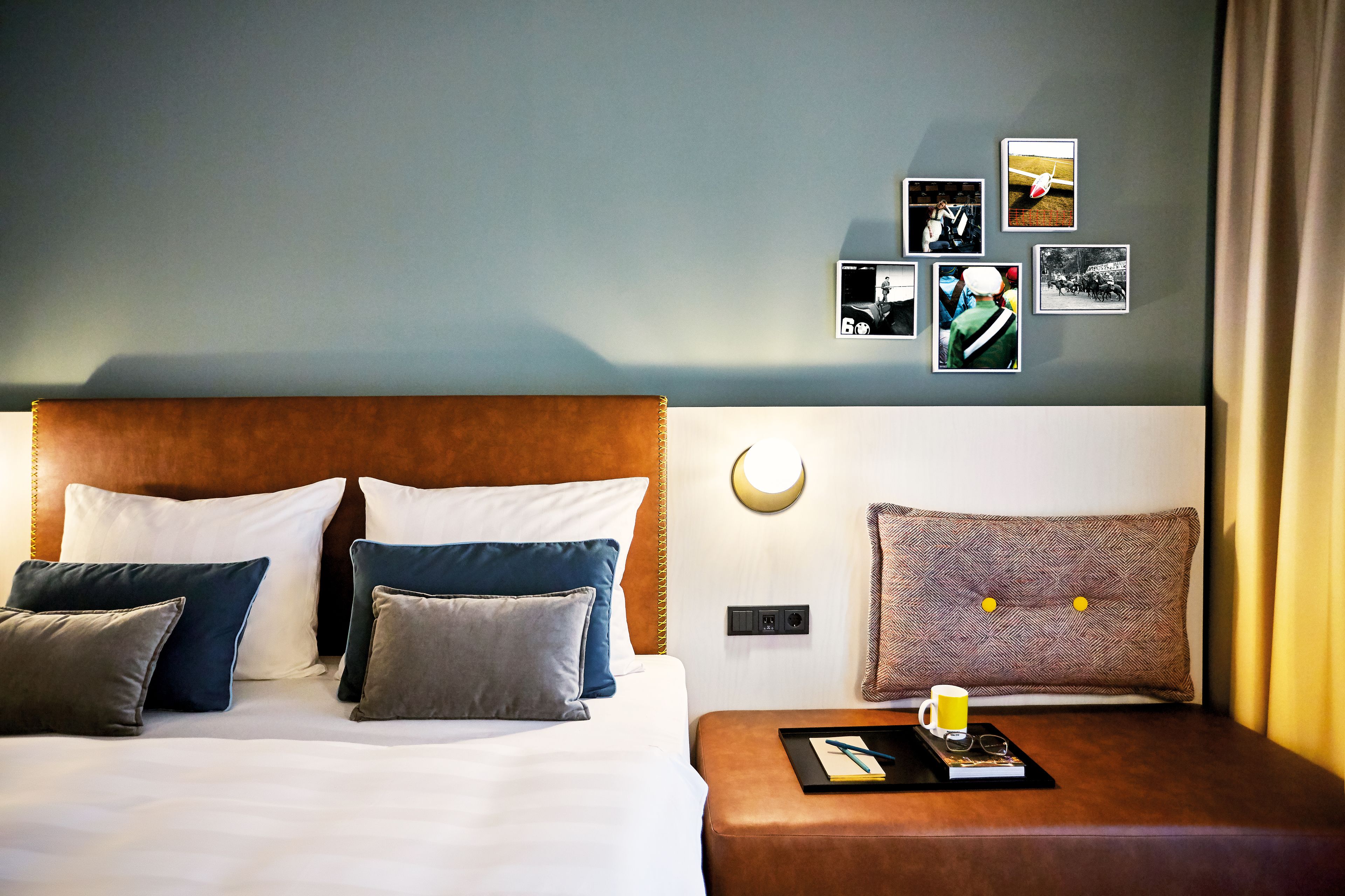 Una cama bien hecha con varias almohadas, una mesita de noche con un libro, gafas y una taza, y una pared con varias fotos enmarcadas.