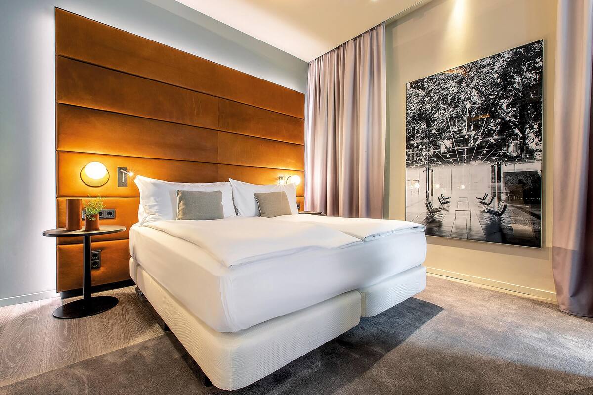 A modern hotel room with a large bed, a nightstand, a small table with a lamp, and a large framed picture on the wall.