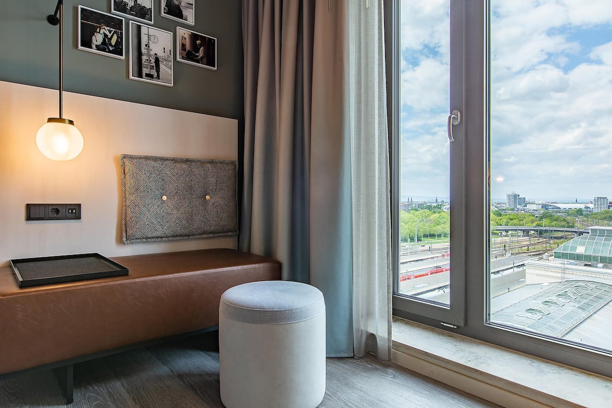 A modern room with a bench, ottoman, wall-mounted light, and a city view through large windows.