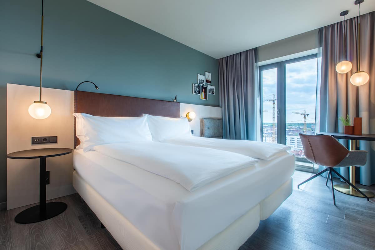 A modern hotel room with a large bed, bedside tables, a desk, and a view of the city.
