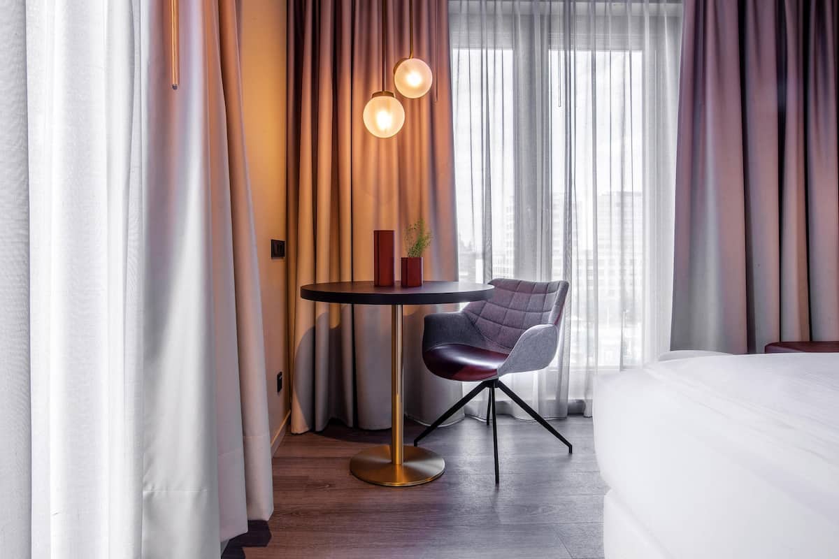 A hotel room with a bed, a round table with a chair, a potted plant, and curtains.