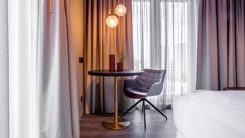 A hotel room with a bed, a round table with a chair, a potted plant, and curtains.