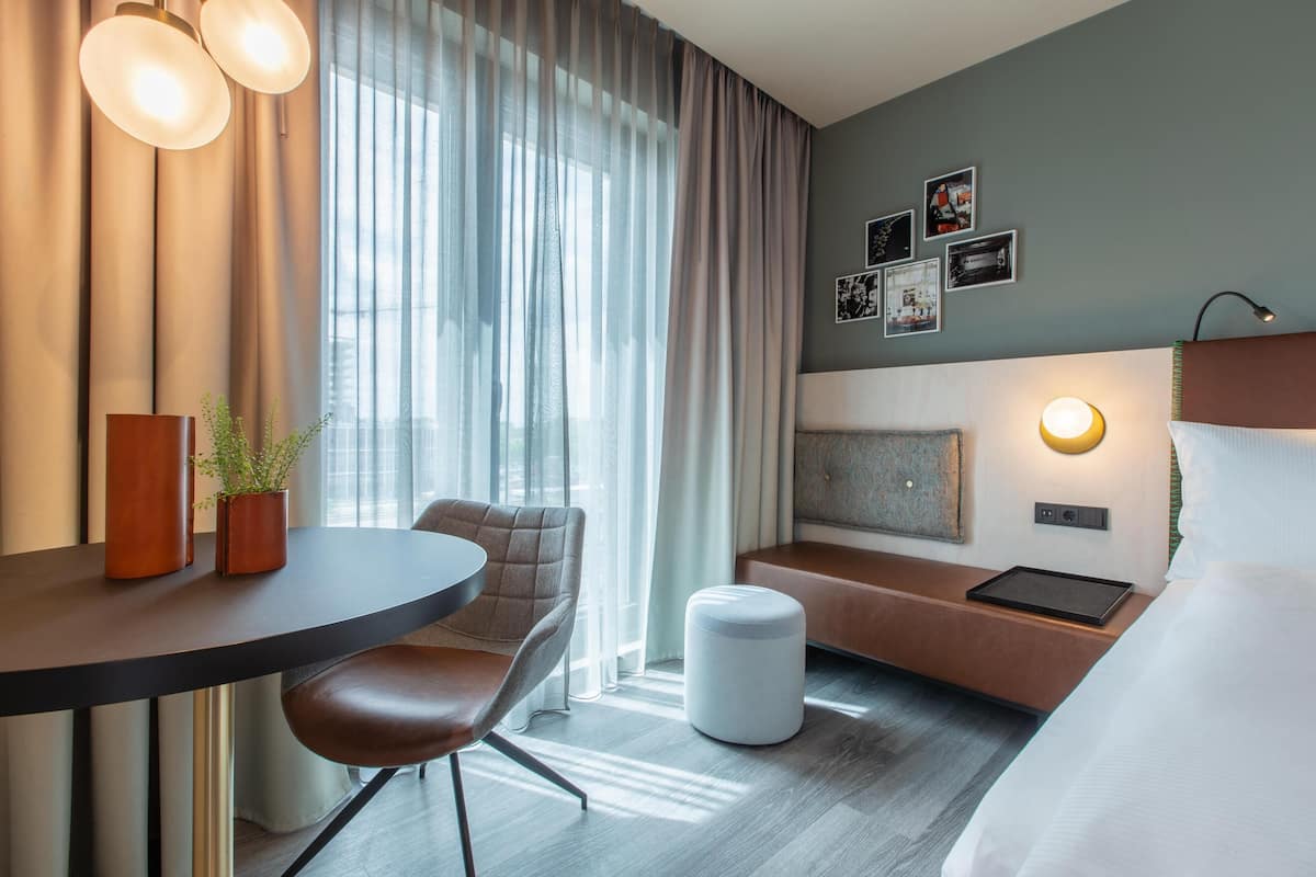A hotel room with a bed, a desk with a chair, a small table with two vases, a bench, and a wall-mounted lamp.