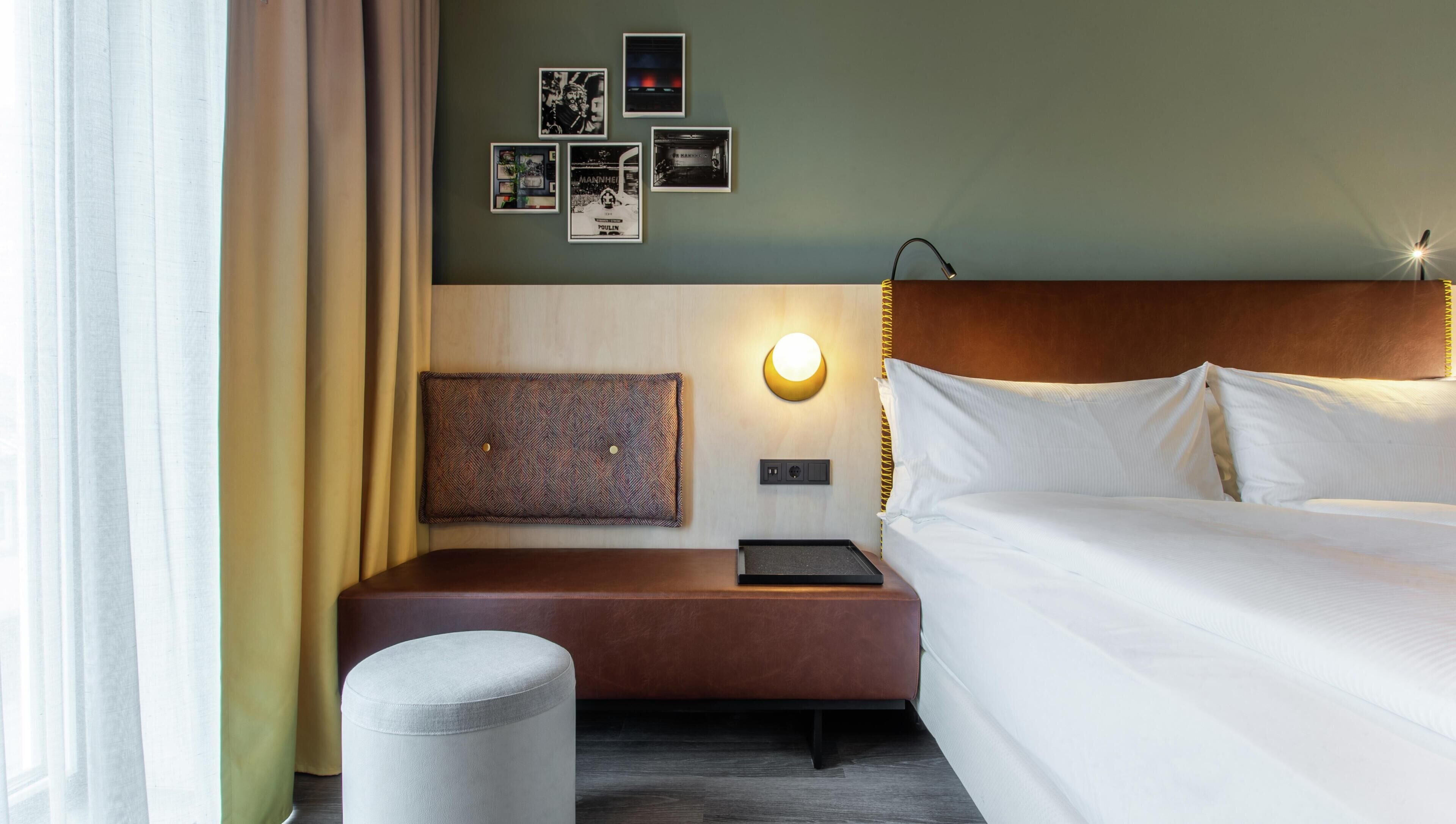 Ein Hotelzimmer mit einem Bett, einem Nachttisch, einer Lampe, einer Wandleuchte, einem gemusterten Kopfteil und einem kleinen Hocker.