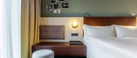 Ein Hotelzimmer mit einem Bett, einem Nachttisch, einer Lampe, einer Wandleuchte, einem gemusterten Kopfteil und einem kleinen Hocker.