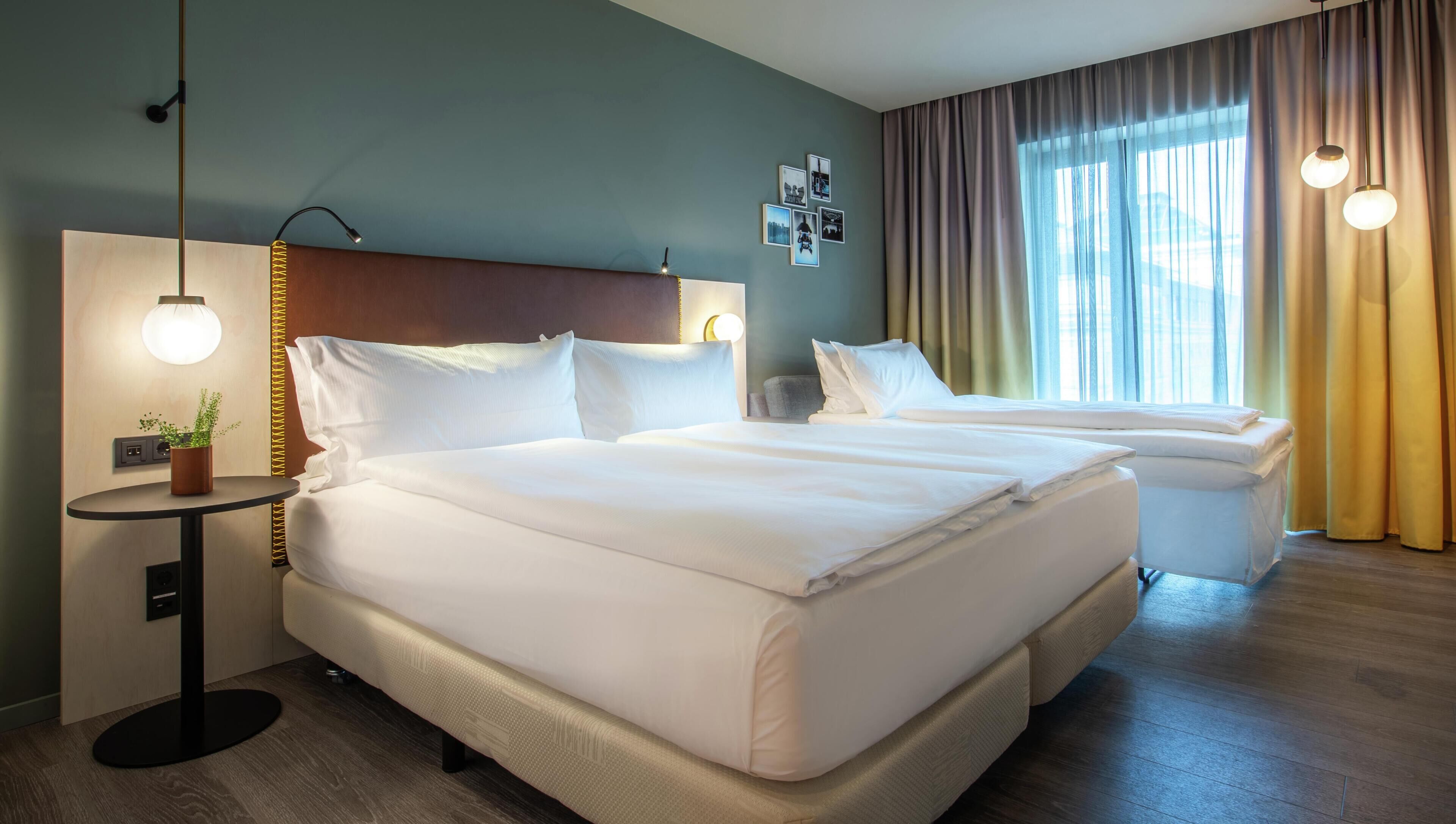 A hotel room with a large bed, a bedside table with a lamp, a small table with a plant, and a window with curtains.