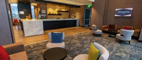 A hotel lobby with a reception desk, seating area, and a Hampton by Hilton sign.
